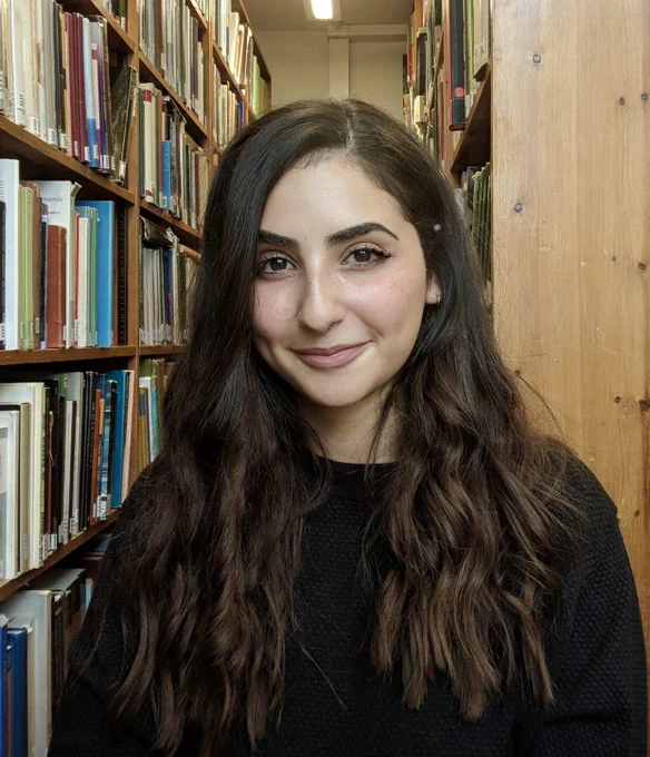 portrait of a person with long dark hair who is standing between book shelves in a library