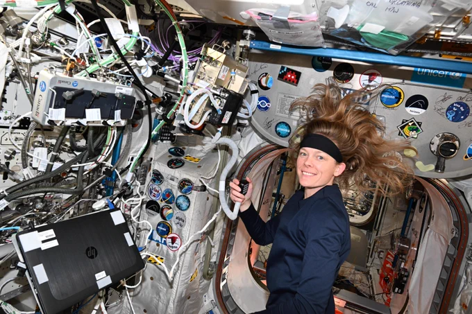 NASA astronaut Nichole Ayers floating inside the space station, holding a radio mic. Her hair fans out in weightlessness as she smiles at the camera. The surrounding area is filled with cables, equipment, and mission patches.