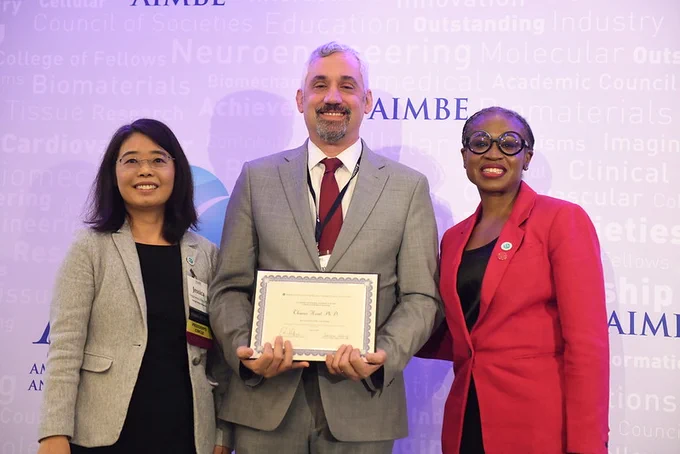 Three individuals stand in front of a conference banner with text related to biomedical engineering. The central person is holding a certificate. All are smiling.