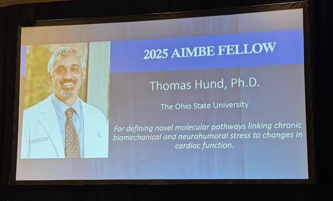 Presentation slide announcing Thomas Hund, Ph.D., as a 2025 AIMBE Fellow from The Ohio State University, for work on molecular pathways related to cardiac function, displayed next to a portrait of a smiling individual in medical attire.