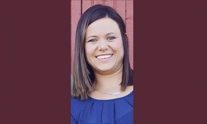 A photo of Ashley on a maroon-colored background. Ashley has brown hair that falls to about her shoulders. She is wearing a silver necklace and has a blue shirt on.