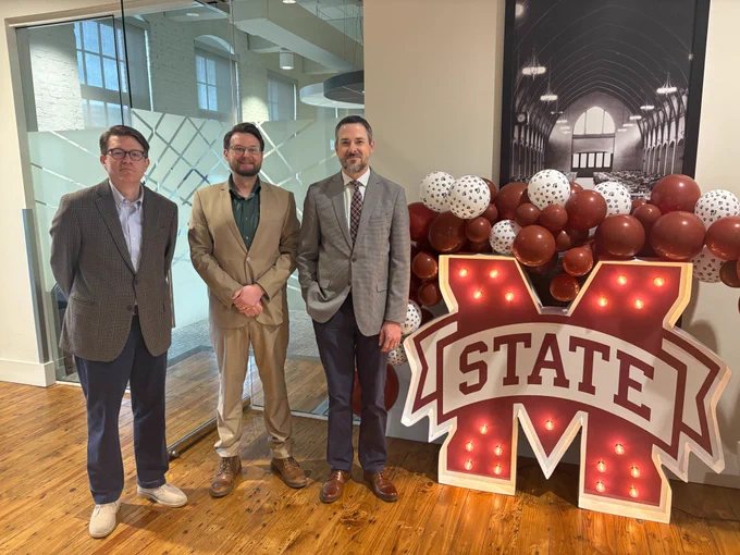 three men in suits standing next to the Mississippi State University "M State" logo