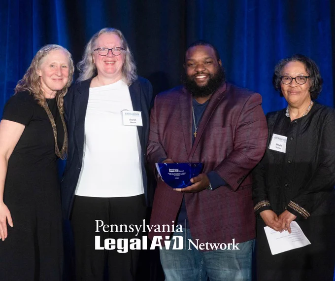 This is an image of PLAN, Inc. Executive Director Meredith Rapkin, Community Legal Services Litigation Director Sharon Dietrich, House Majority Appropriations Chairman Jordan Harris, and MidPenn Legal Services Executive Director Rhodia Thomas standing together at the 2025 Excellence Awards against a blue backdrop. Chairman Harris, who delivered the keynote address, holds a blue bowl that was given to him as a token of appreciation. The PLAN, Inc. logo appears at the bottom of the image.