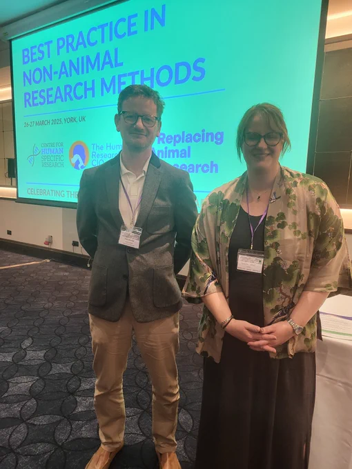 Dr. Owen Kavanagh and Celean Camp stand side-by-side in front of a projected screen with the title of the conference, "Best Practice in Non-Animal Research Methods"