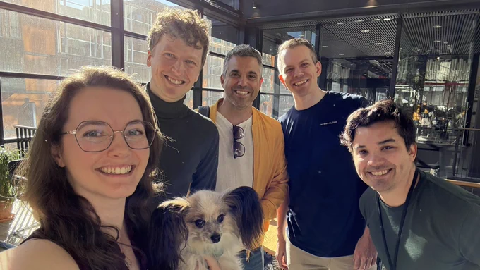Group photo of Sofia, Anton, Daniel, Oscar, César and Ellie (a papillon dog), all smiling to the camera. The sun shines in from behind through the big glass outer walls of WeWork.