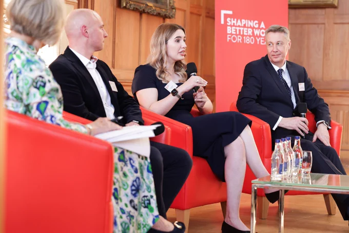 Image shows Claire Wilson, KTP associate speaking while on a panel. The other panel members are listening to her speak. They are sitting on red tub chairs with a red Queen's University Belfast banner behind them. In front of them is a table with bottles of water on it.