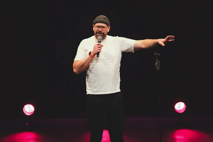 A male comedian wearing glasses with a dark beanie hat. He has a white t-shirt on and he is gesturing with his arm outwards. The stage is dark and dimly lit.