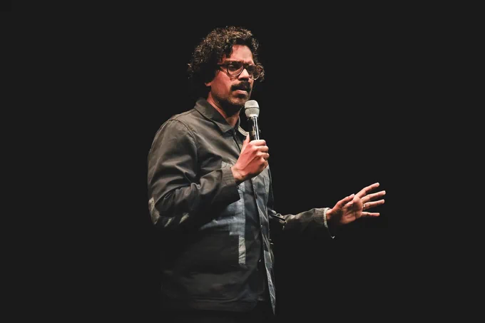 A male comedian with dark curly hair wearing a black jacket and wearing glasses. He is holding a microphone. The background is dark and the stage is lit.