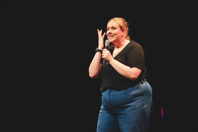 A female comedian with light skin and blonde hair tied back is performing on stage. She is wearing a black short-sleeved top and blue jeans. Holding a microphone in her right hand, she gestures with her left while smiling. The background is dark, with a spotlight illuminating her.
