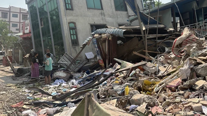 Three people stand amidst debris and rubble in a street with destroyed and collapsed buildings.