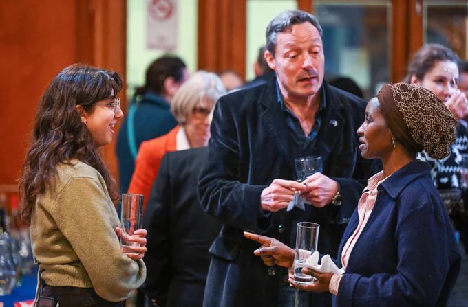 Three people talking to each other with drinks in hands. They're inside a church and surrounded by other people of different ages.