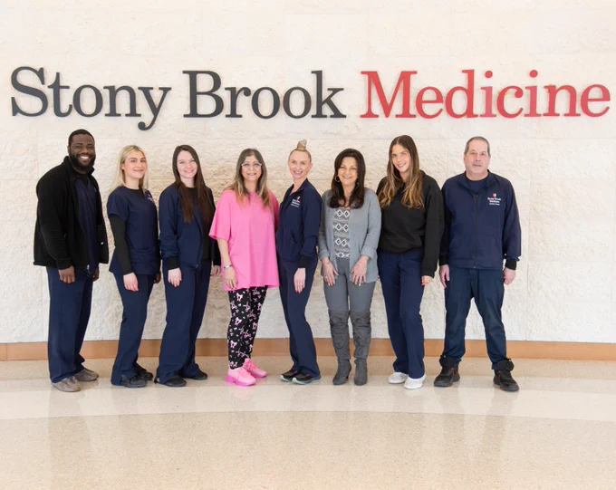 Group of eight medical staff members smiling and standing in front of a wall with the Stony Brook Medicine logo.