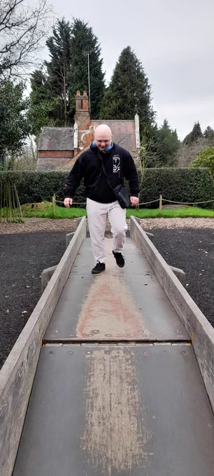 a man walks along a wooden walk way. He's wearing a black sweatshirt and cream trousers. Behind him is the roof of a house, the rest of the house is hidden by a green hedge. 