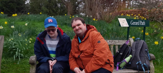 Two men sitting on a bench outdoors. Behind them is lots of green grass, yellow daffodils and a hedgerow. There are rucksacks next to the men on the bench. 