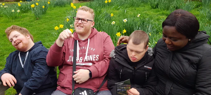 A group of three men and one woman sitting on a bench outdoors. Behind them is bright green grass and lots of clumps of yellow daffodils. 