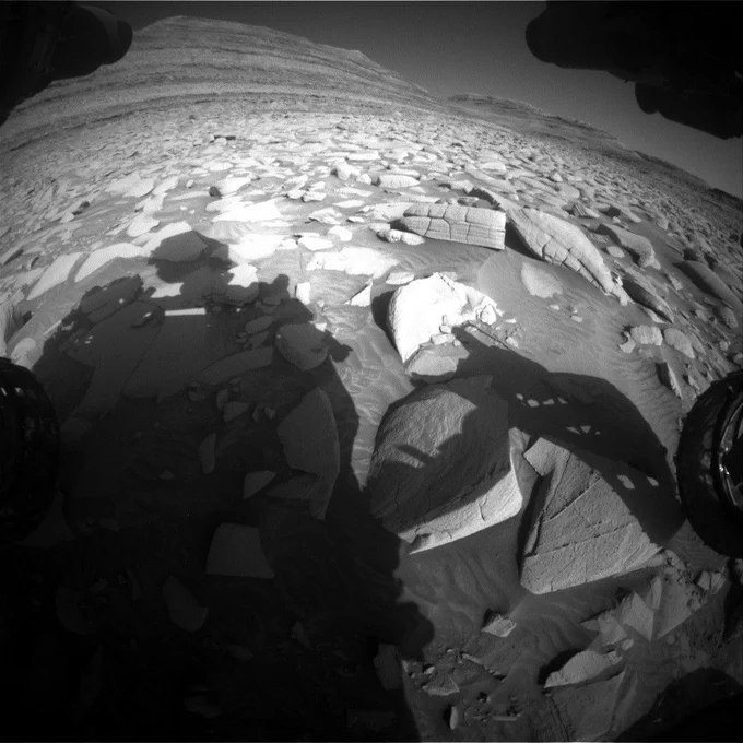 A black-and-white image from the Curiosity rover shows a rocky, uneven terrain with scattered flat, angular stones on Mars. In the foreground, the rover's dark shadow stretches across the light-colored, dusty Martian surface. The shadow of the robotic arm is visible, extending toward a large, cracked rock. The background features a sloping, layered hill with horizontal striations, indicating sedimentary rock formations. The sky appears dark, contrasting with the sunlit landscape.
