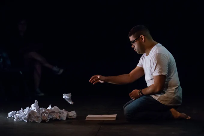 Photo by Paulina Kim Joo.
 
Description
 
In a darkened space, a performer kneels in front of a pile of crumbled white paper. He is in the motion of throwing a new crumbled paper into the pile.