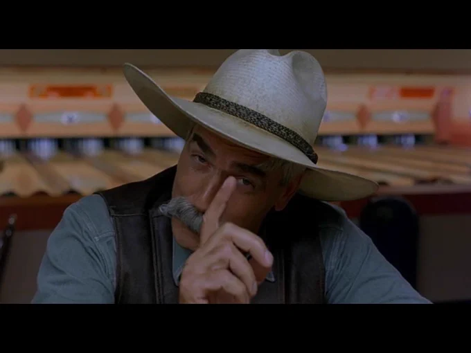 The Stranger cocks his head and gestures with a raised index finger towards the camera.