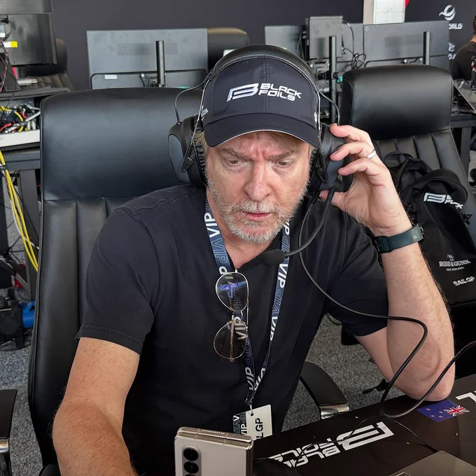 Picture of Rhys Darby in a black short sleeved shirt with a Black Foils hat on, wearing a headset and holding it up to his ear. He's looking down at a screen and a phone intensely.