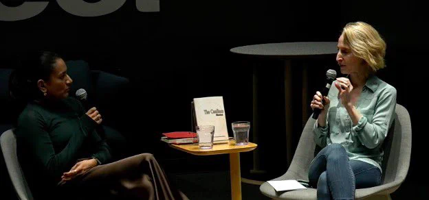 Author Anu Kandikuppa is wearing a long sleeve, dark green shirt and silk pants.  Her book THE CONFINES is sitting on a table. Across from her is Michelle Hoover. Michelle is wearing blue jeans and a light green button up shirt. They both have microphones and are sitting on a lighted stage.