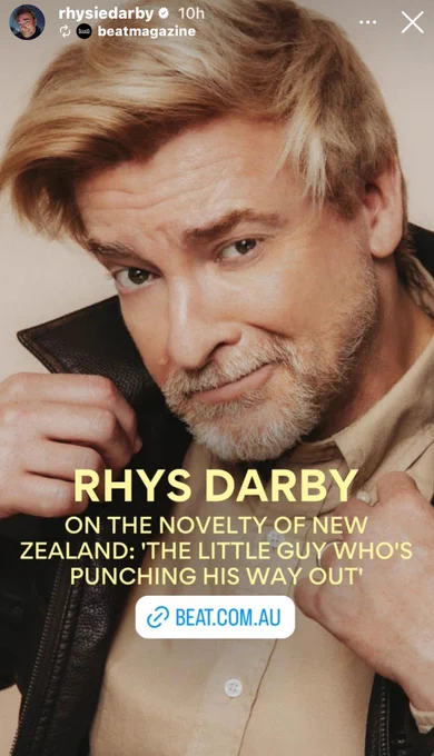 Picture of Rhys Darby holding his jacket lapels up. Yellow text states Rhys Darby On the Novelty of New Zealand: ' The Little Guy Who's Punching His Way Out' - link to be beat.com.au
