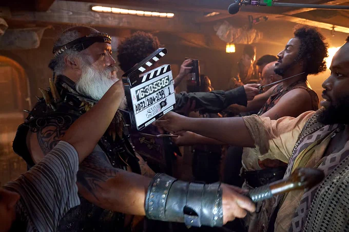 David Fane, as Fang, Samson Kayo as Oluwande hold weapons to each other with other crew members in the background doing the same, a set hand holds up the marker for filming