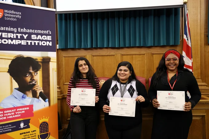 Three individuals holding certificates stand in front of a banner for Middlesex University London's "Diversity SAGE Competition," with a promotional poster featuring a character on the left side.