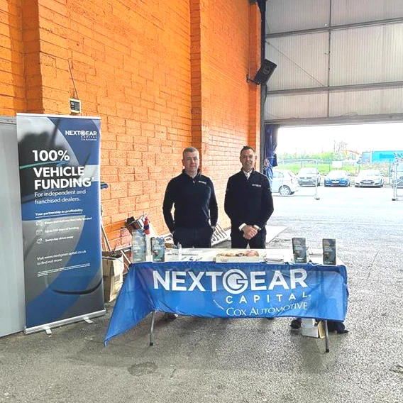 Two individuals standing behind a promotional booth for NextGear Capital at an event, with banners and merchandise on display.