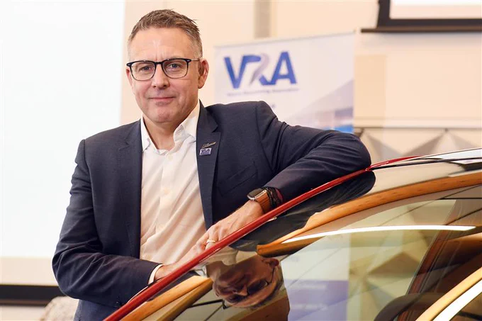 Philip Nothard in a business suit standing beside a car indoors, with a sign displaying "VRA" in the background.