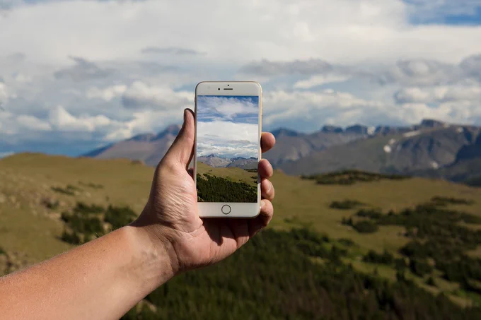 A person holds a mobile phone out at arms length to take a picture of a mountain range in the distance under a cloud-filled sky