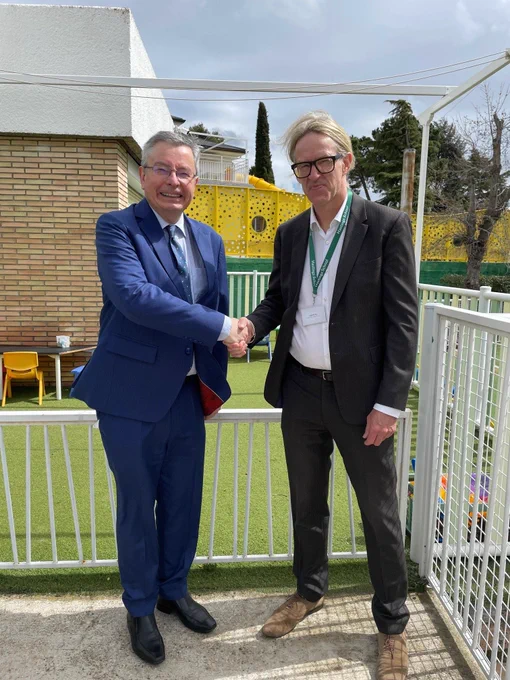 Two men in business attire shake hands outdoors on a fenced playground area. The man on the left wears a blue suit and tie, while the man on the right wears a dark suit with a green lanyard. In the background, there is artificial grass, a yellow play structure, and trees.