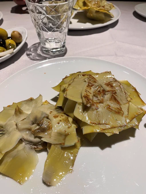 Plate of grilled or sautéed artichoke slices arranged in a fan-like pattern, lightly browned and glistening with oil. In the background, a glass of water and a bowl of assorted olives rest on a white tablecloth.