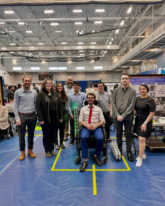A group photo of an engineering capstone group with their project, an accessibility solution for railway transit. The student in the centre of the group photo is seated in the wheelchair that is being used for a project demonstration.