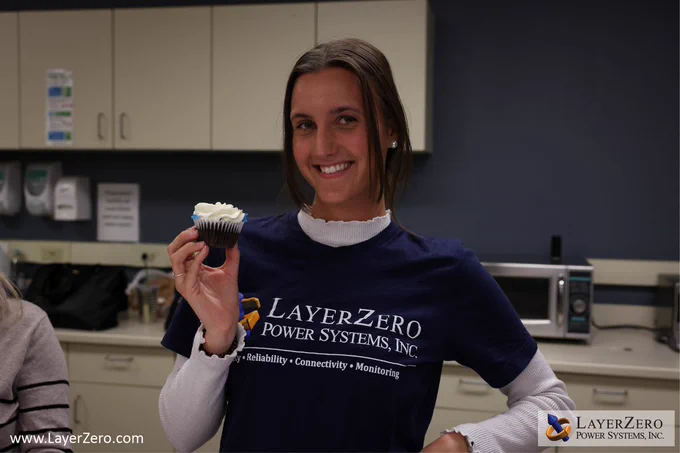 A female employee with brown hair smiles while holding a chocolate cupcake with white frosting. She is wearing the LayerZero logo on her shirt. The bottom left corner has text that reads www.LayerZero.com. The bottom right corner has the LayerZero logo with a white box behind it.