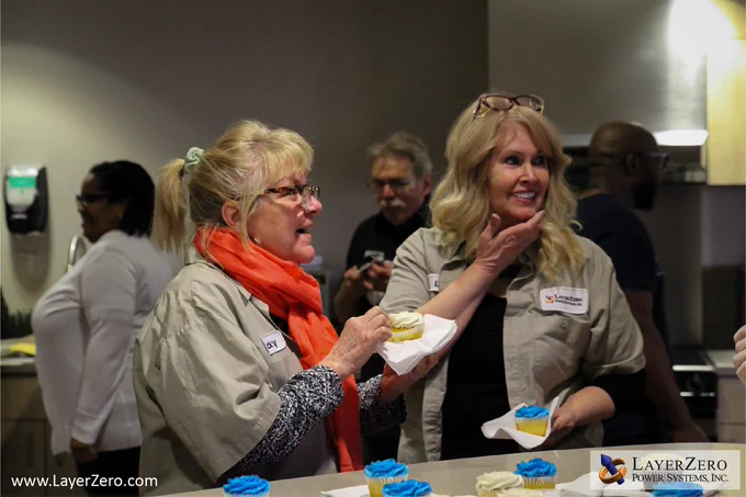 A pair of two female employees with blonde hair smile at the person serving cupcakes off screen. The employee on the left is holding a vanilla cupcake with white frosting and the employee on the right is holding a vanilla cupcake with blue frosting. There are three employees visible in the back of the photo. The bottom left corner has text that reads www.LayerZero.com. The bottom right corner has the LayerZero logo with a white box behind it.