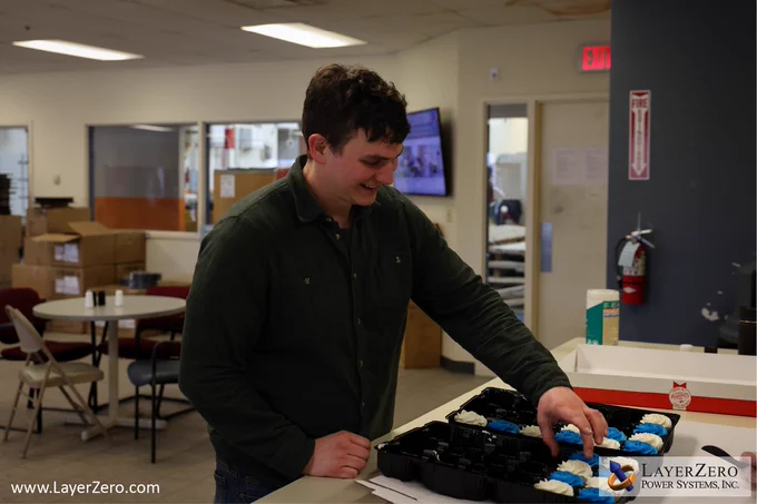 A male employee wearing a long sleeves green shirt reaches for the tray of cupcakes. He is smiling as he grabs a chocolate cupcake with white frosting. The bottom left corner has text that reads www.LayerZero.com. The bottom right corner has the LayerZero logo with a white box behind it.