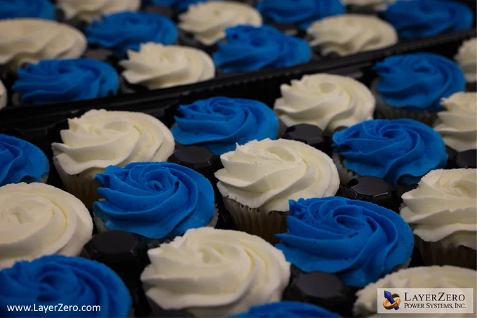 The image shows a tray of chocolate and vanilla cupcakes with alternating white and blue frosting. The bottom left corner has text that reads www.LayerZero.com. The bottom right corner has the LayerZero logo with a white box behind it.