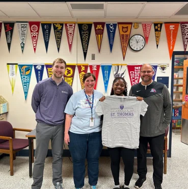 Four adults in a classroom displaying university pennants on the wall, with one person holding a St. Thomas t-shirt. They are smiling and standing closely together.