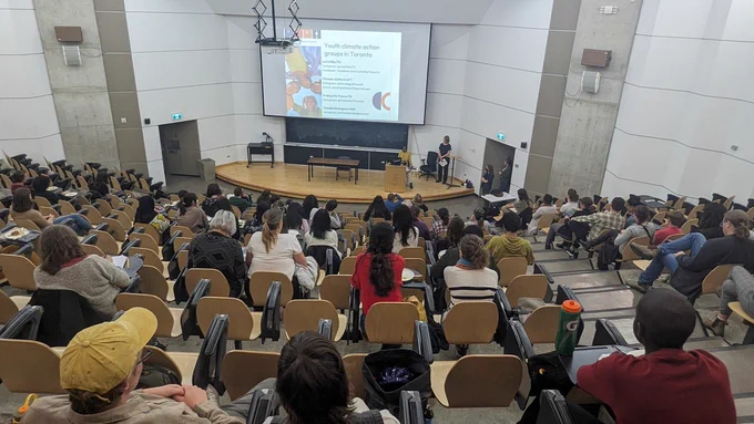 Many students sit in a big university lecture hall with a sunken stage while someone gives a presentation. 
