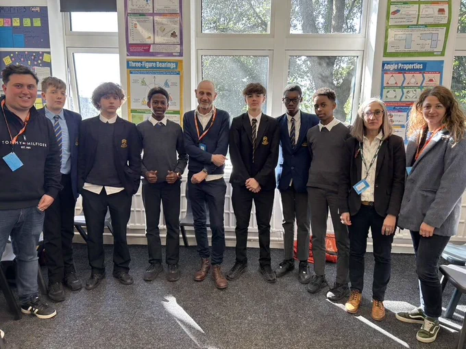 A group of students and adults standing together in a classroom. Five secondary school boys in uniform stand in the center, flanked by four adults and two other students. 