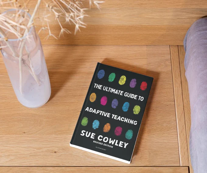 Picture of a copy of The Ultimate Guide to Adaptive Teaching on a wooden desk next to a vase of dried flowers. The book has a black cover with colourful fingerprints.