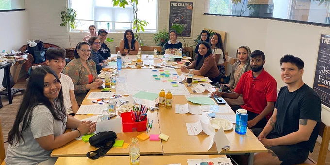 A group of students sit around a large table with many papers and work supplies throw across the table. 