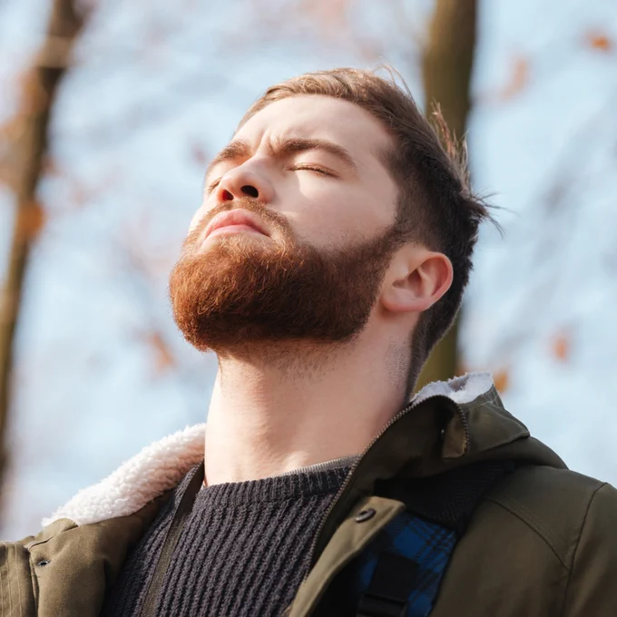 A bearded man wearing a green jacket closes his eyes and tilts his head upward while taking a deep breath in nature.