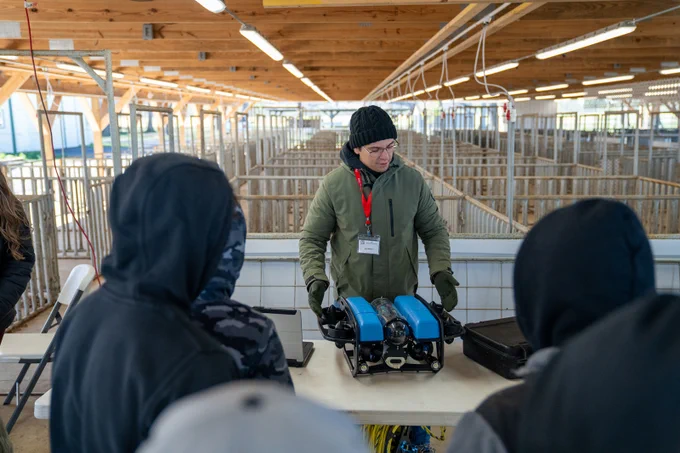 A scientist standing behind a folding table shows 7th grade students a small, underwater robot used for oyster aquaculture research