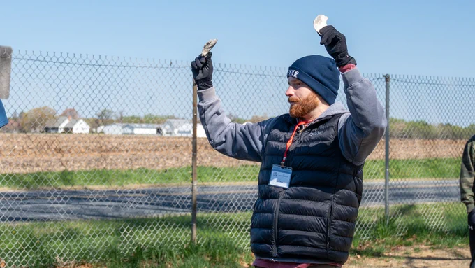 An educator standing outdoors near a farm and a wire fence holds up two oyster shells while talking to students