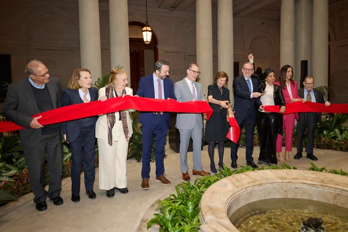 Nine individuals lined up holding a bright red ribbon in a garden court
