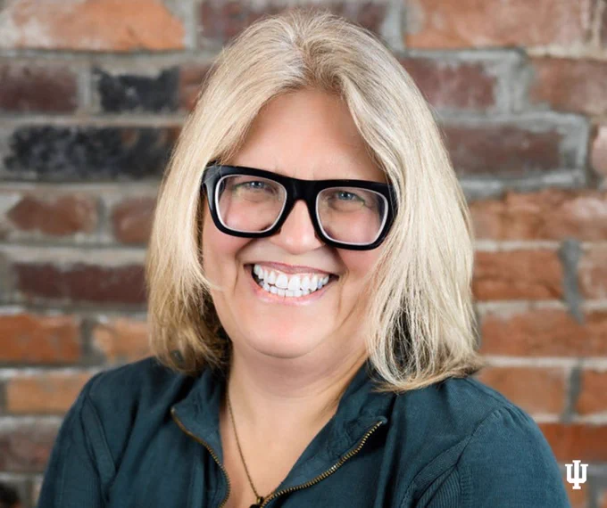 Portrait of a smiling person with blonde hair wearing glasses, in front of a brick wall with the Indiana University logo in the corner.