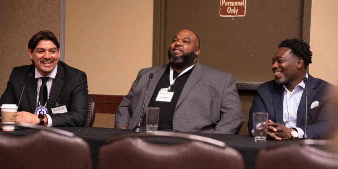 Three individuals sitting at a panel table, smiling and engaged in a discussion at a conference room. There is a "Personnel Only" sign on a door in the background.
