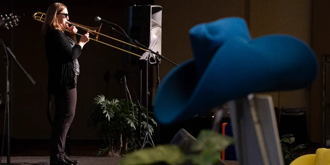 Person playing a trombone on stage at a conference, with a large blue hat in the foreground.