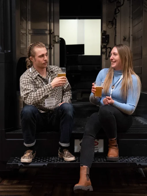 Two individuals smiling and conversing while holding drinks, seated on a step.